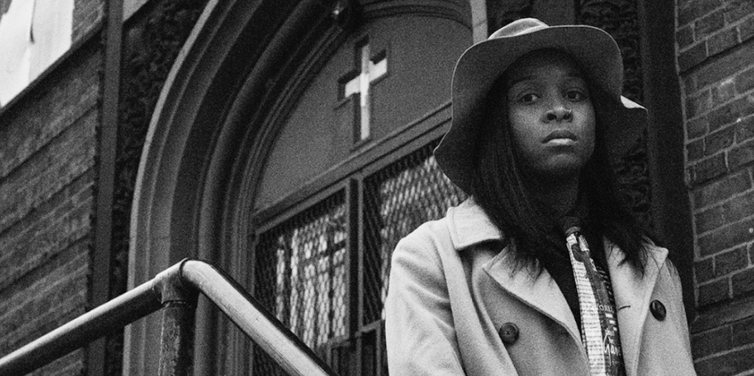 Black woman wearing hat stands in front of a building with a cross on the front. photo in black and white